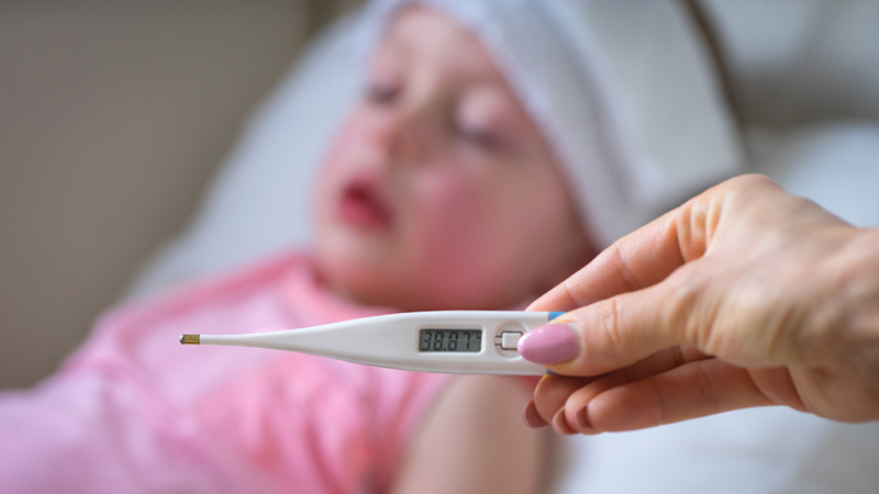 criança pequena com aspeto adoentado e mão feminina com termómetro digital